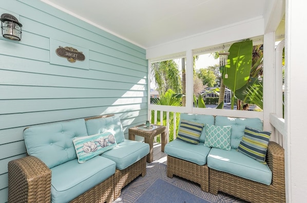 A covered porch with two light blue cushioned sofas and wicker furniture, a small table, and a sign on the wall.