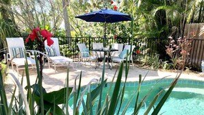 A pool area with lounge chairs and a table, surrounded by tropical plants and a wooden fence. - Sunset Beach 2 Bedroom Villa by RedAwning (Holmes Beach)