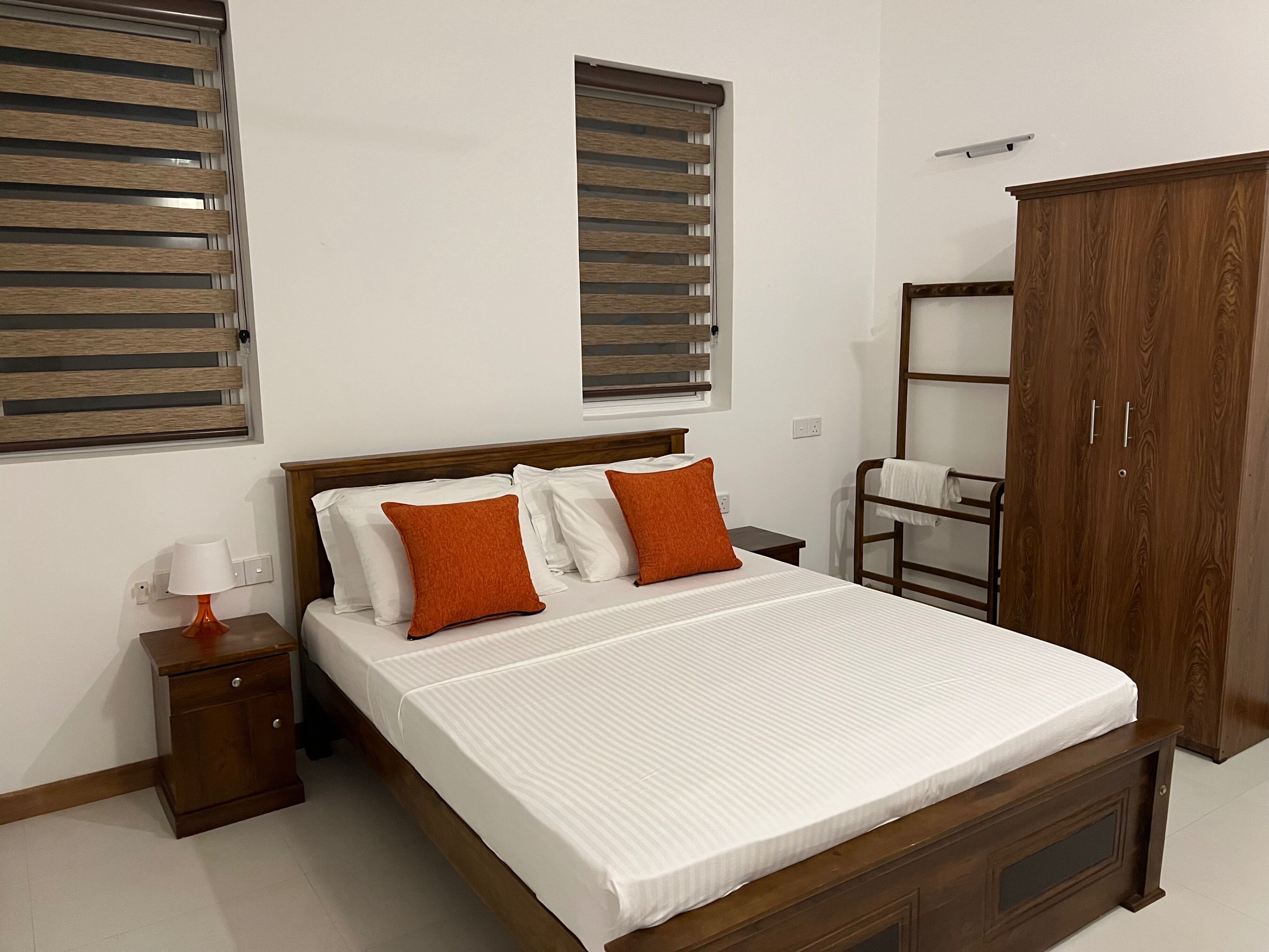 Un dormitorio con una cama de madera, dos ventanas con persianas, un armario, una mesita de noche con una lámpara y un escritorio pequeño.