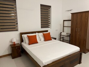 A bedroom with a wooden bed, two windows with blinds, a wardrobe, a bedside table with a lamp, and a small desk. - Silina Airport Residence in Katunayake (Seeduwa - Katunayake)