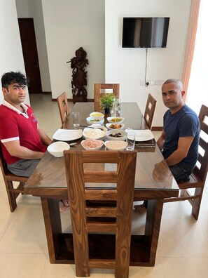 Two people seated at a wooden dining table with various dishes, a potted plant, and a TV mounted on the wall. - Silina Airport Residence in Katunayake (Seeduwa - Katunayake)