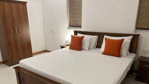 A neatly made bed with white linens and two orange pillows, a wooden headboard, and a nightstand with a lamp. - Silina Airport Residence in Katunayake (Seeduwa - Katunayake)