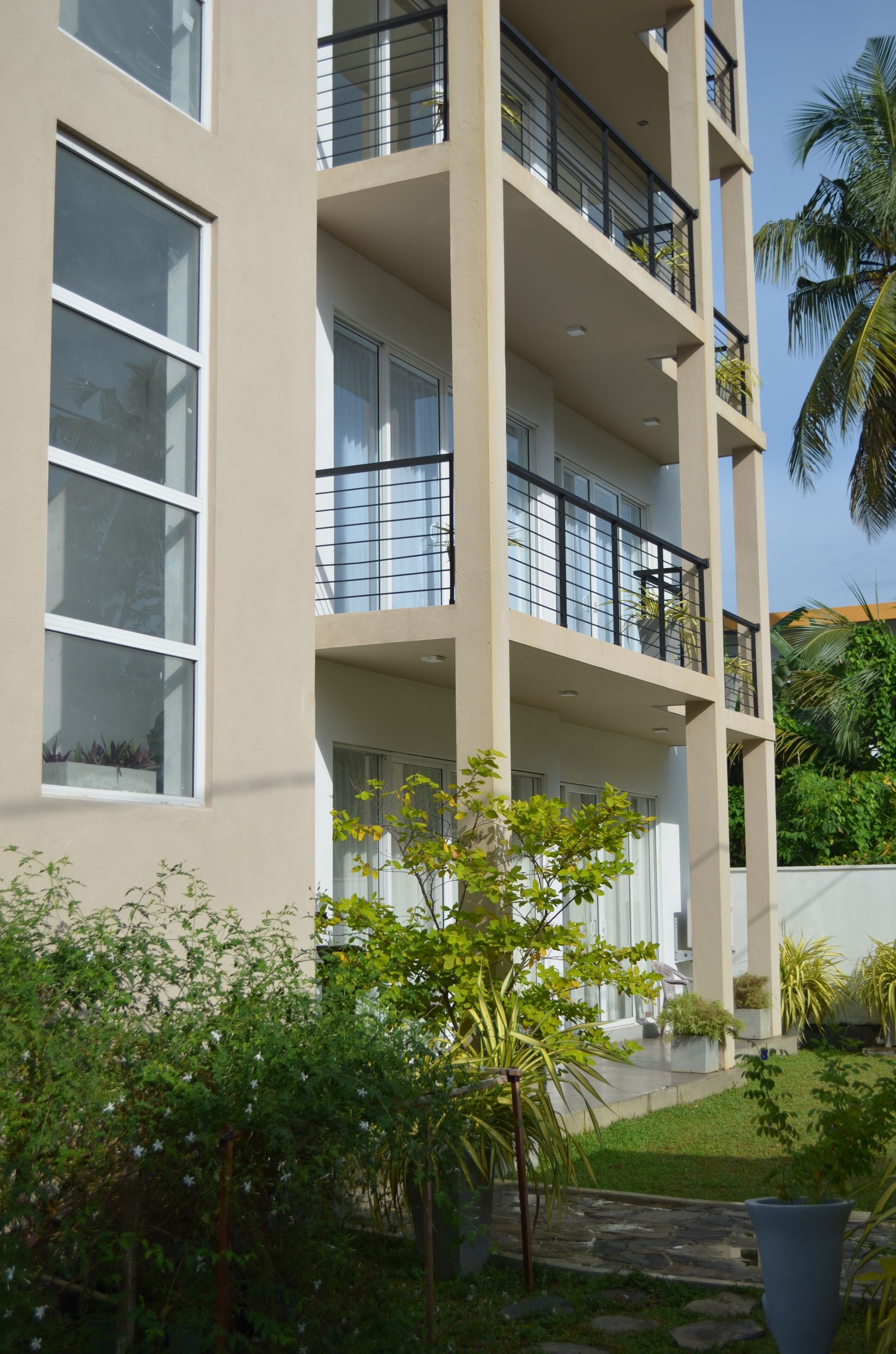 A multi-story building with balconies, large windows, and a garden area with various plants.