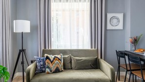 Ein Wohnzimmer mit einer Couch, einer Stehlampe, einem Esstisch mit Stühlen und einem Fenster mit Vorhängen.