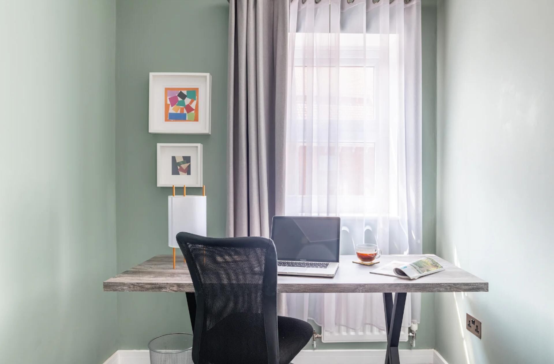 A room with a desk, a chair, a laptop, a glass of tea, and two framed artworks on the wall.