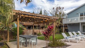 A wooden pergola with string lights, a patio table with chairs, and a poolside area with lounge chairs.