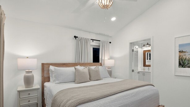 A bedroom with a wooden bed, white bedding, two bedside tables with lamps, a ceiling light, and a door leading to a bathroom.
