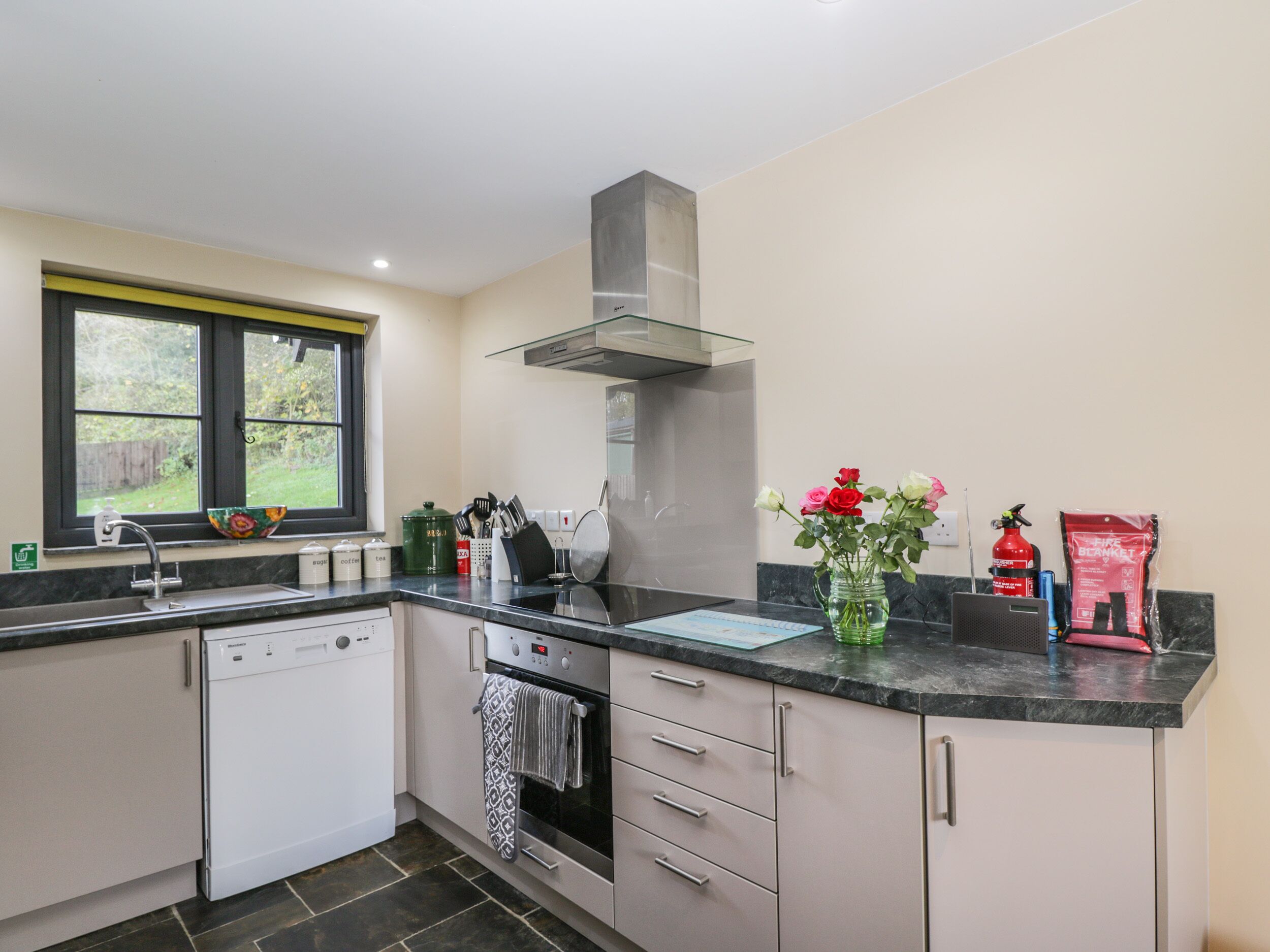 A modern kitchen with a dishwasher, oven, and a window overlooking a garden.