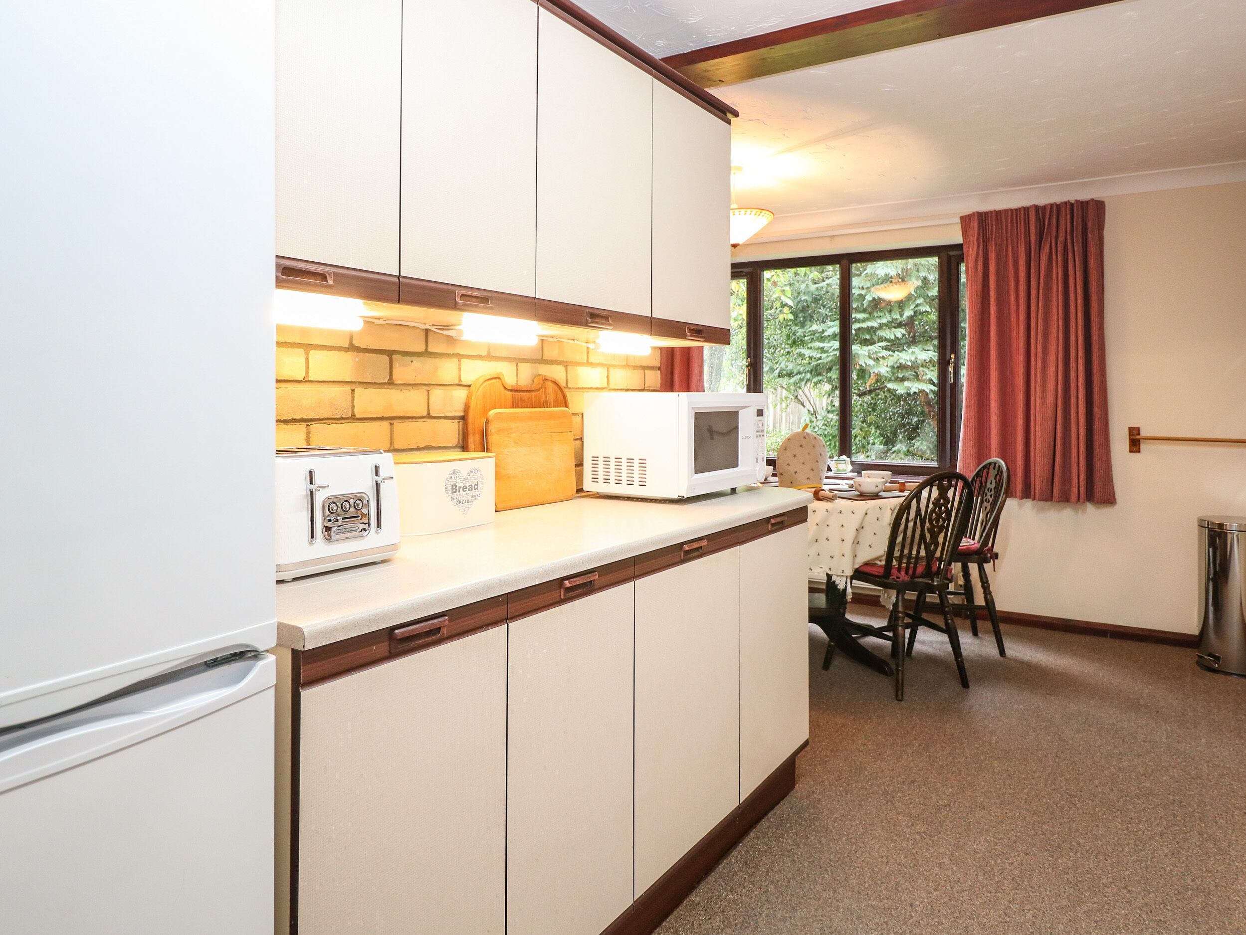 A compact kitchen with white cabinets, a microwave, a toaster, and a bread box. There is a dining table with chairs and a window with curtains.