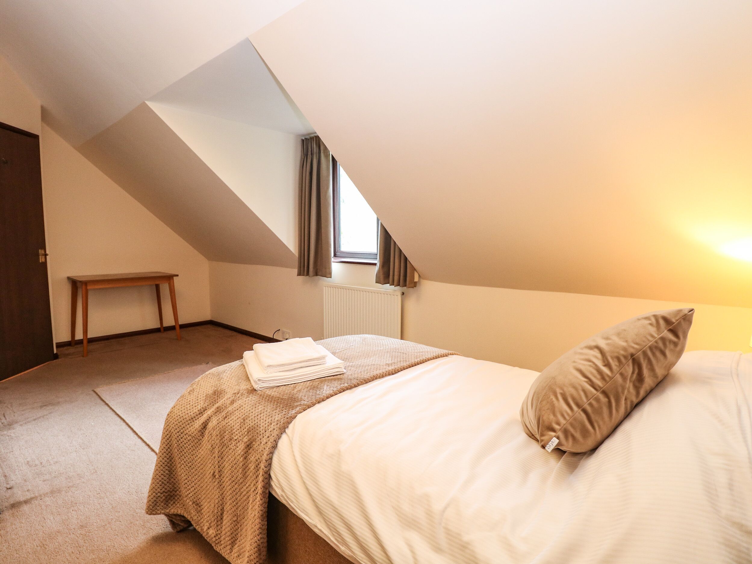 Une chambre avec un plafond en pente, un lit recouvert d’une couverture et d’un oreiller beiges, une petite table en bois et une fenêtre avec des rideaux.