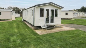 A single mobile home with a small porch and a chair, surrounded by a grassy area and other similar homes in the background. - Pines 30 (Scarborough)