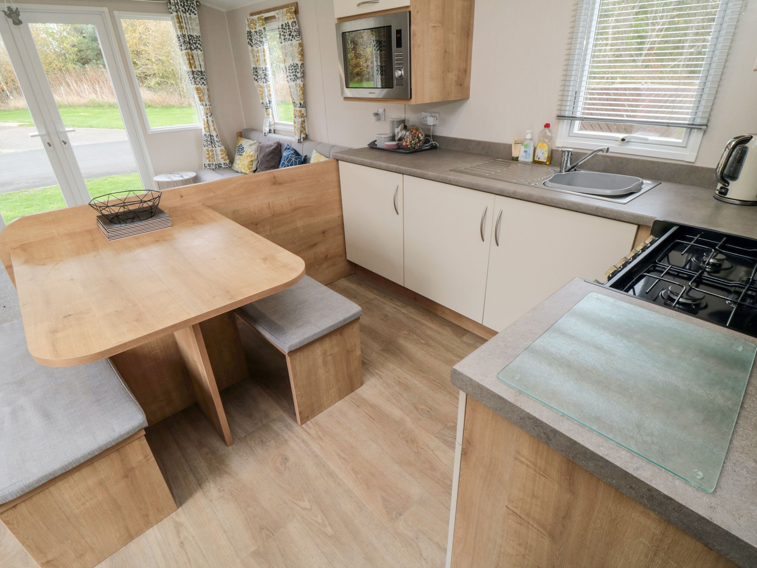 A compact kitchen with a wooden table, bench seating, a built-in oven, and a window with blinds.