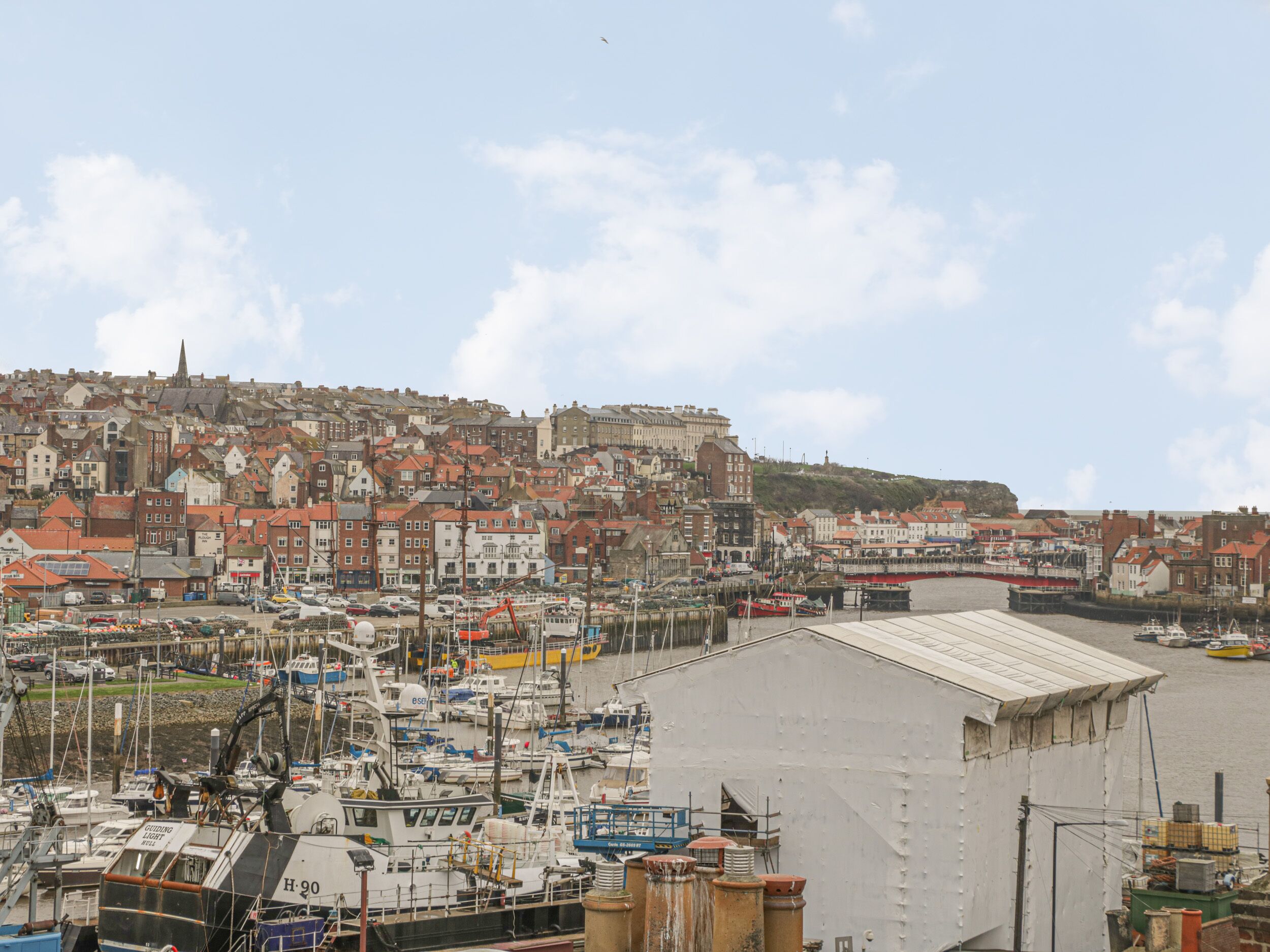 A harbor with numerous boats docked, a large industrial building, and a densely packed town with historic buildings in the background.