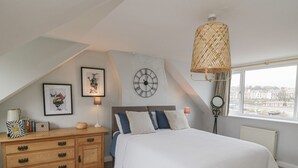 A bedroom with a bed, a wooden dresser, a wall clock, and a window with a view of buildings.