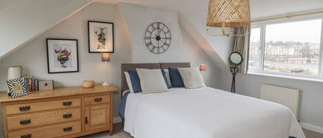 A bedroom with a bed, a wooden dresser, a wall clock, and a window with a view of buildings.