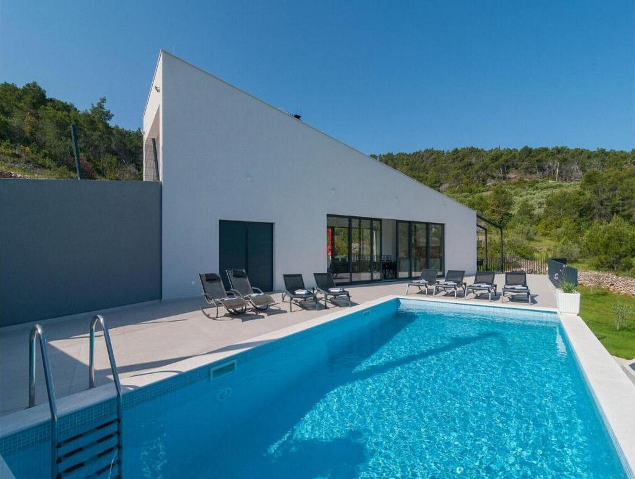 Un área moderna junto a la piscina con una piscina azul cristalina, sillas de descanso y un edificio blanco con ventanas amplias.