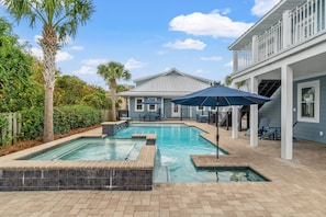 A swimming pool with a stone border, surrounded by a patio area with chairs and umbrellas.
