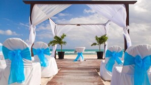 Une jetée en bois mène à un espace aménagé pour un mariage sur la plage, avec des chaises blanches et des rubans bleus, ainsi que deux palmiers en pot.