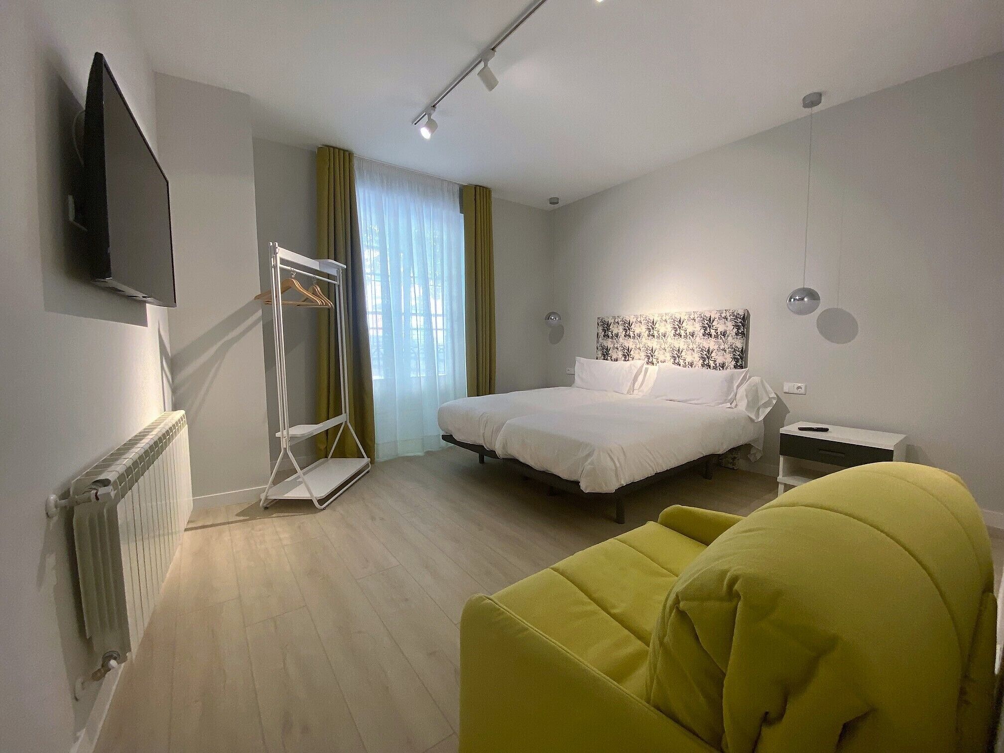Habitación de hotel con cama, televisor montado en la pared, radiador, ventana con cortinas y una silla amarilla.