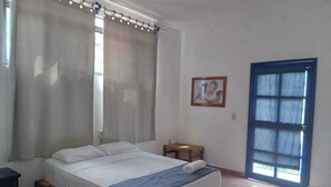 Une chambre à coucher avec un lit, des rideaux, une petite table, une chaise, une porte et un tableau encadré au mur.
