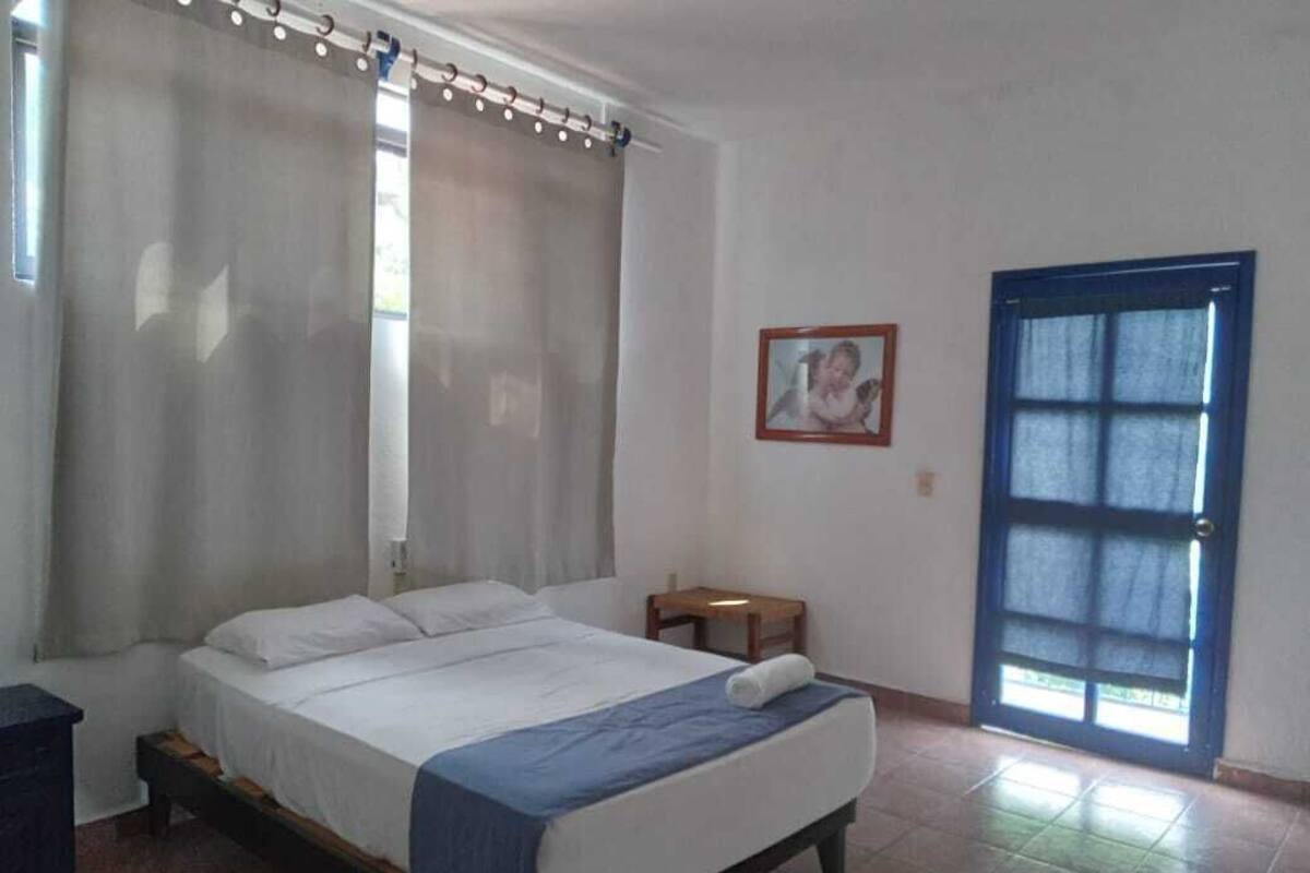Ein Schlafzimmer mit Bett, Vorhängen, einem kleinen Tisch, einem Sessel, einer Tür und einem gerahmten Bild an der Wand.