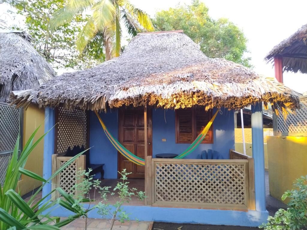Una casa con techo de paja, una hamaca, valla de celosía de madera y pared azul.