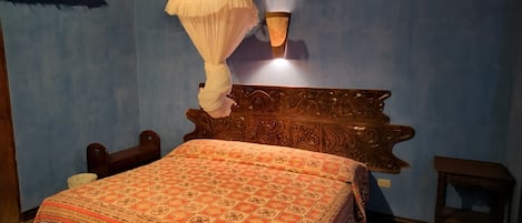 A bedroom with a bed, a nightstand, a bedside table, a hanging mosquito net, and a wall-mounted lamp.