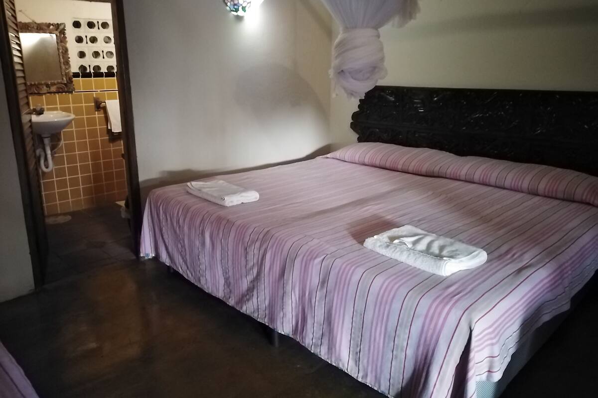 A room with a bed, a bedside table, a hanging light fixture, and a bathroom with a sink and mirror.