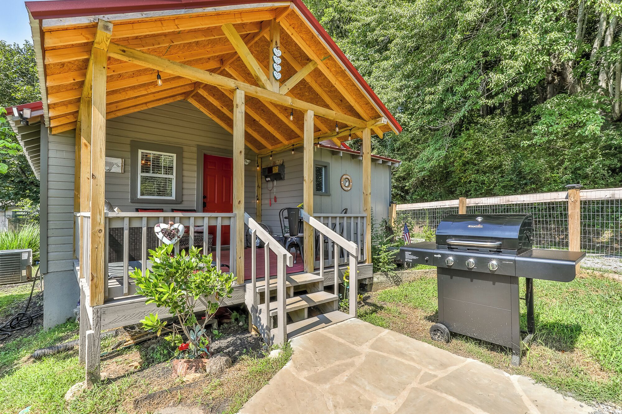 A small wooden cabin with a porch, a barbecue grill, and a red door.
