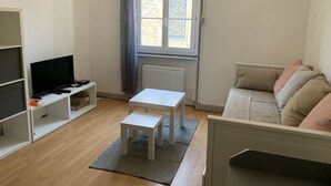 Huoneessa on jalustalla oleva televisio, pieni valkoinen pöytä, koristetyynyillä varustettu sohva ja ikkuna, jossa on verhot.