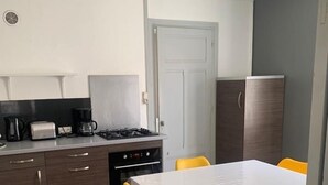 Kompakti keittiö, jossa on valkoinen pöytä ja keltaiset tuolit, sisäänrakennettu uuni ja kahvinkeitin.