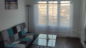 Een woonkamer met een bank, een salontafel en uitzicht op gebouwen door een raam met dunne gordijnen.