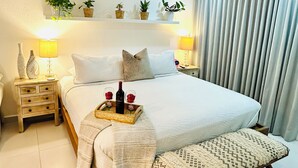 A neatly arranged bedroom with a bed, bedside tables, a bedside lamp, a tray with wine and roses, and a shelf with potted plants. - 5 minutes to SJU airport, Steps to Isla Verde Beach, 16th Fl. w/Prkg @ESJ Towers (Carolina)