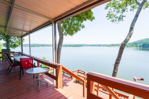 A wooden deck with a table and chairs, overlooking a calm lake with trees and hills in the background.