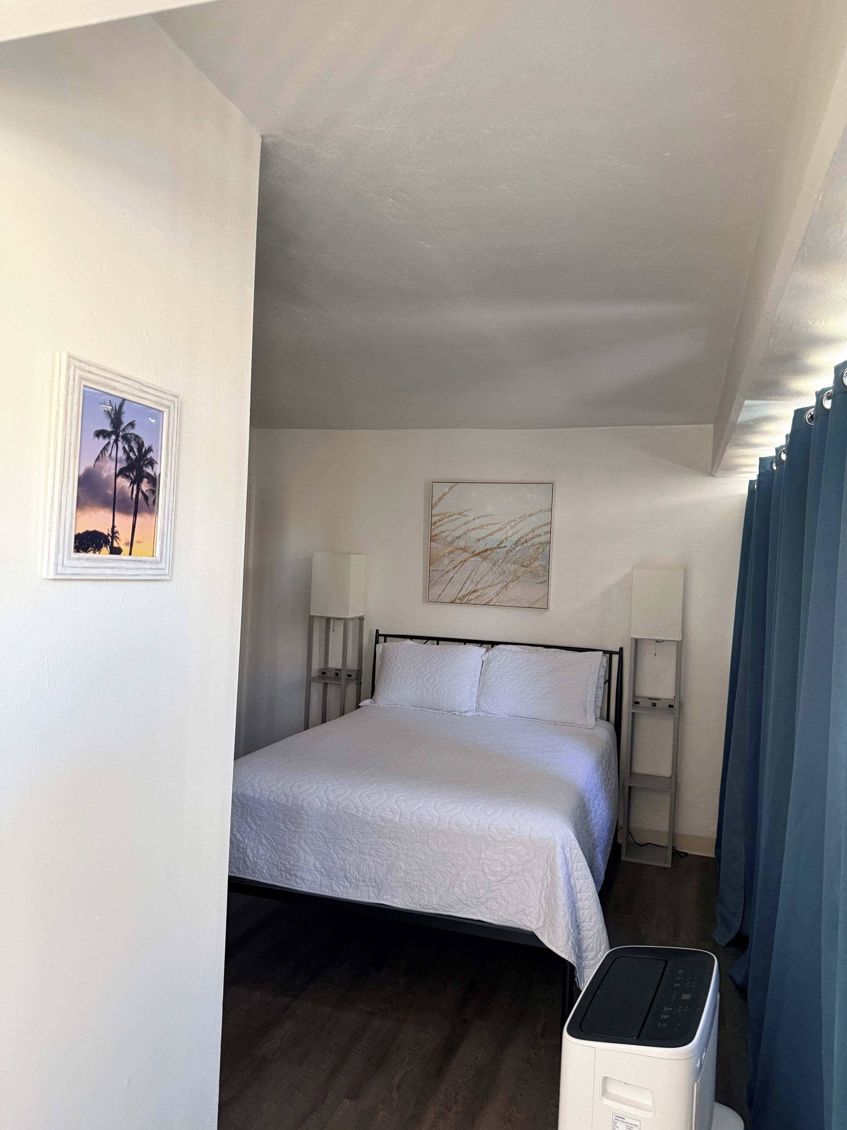 Hotelkamer met een bed, twee bedlampen, een plafondventilator, een foto van palmbomen en een schilderij van abstracte lijnen.