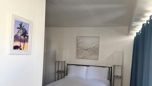 Quarto de hotel com cama, duas luminárias laterais, ventilador de teto, foto de palmeiras e pintura de linhas abstratas.