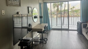 Uma sala de estar moderna com uma grande porta de vidro deslizante que dá para uma varanda, uma escrivaninha com um computador, uma cadeira e uma pequena lixeira.