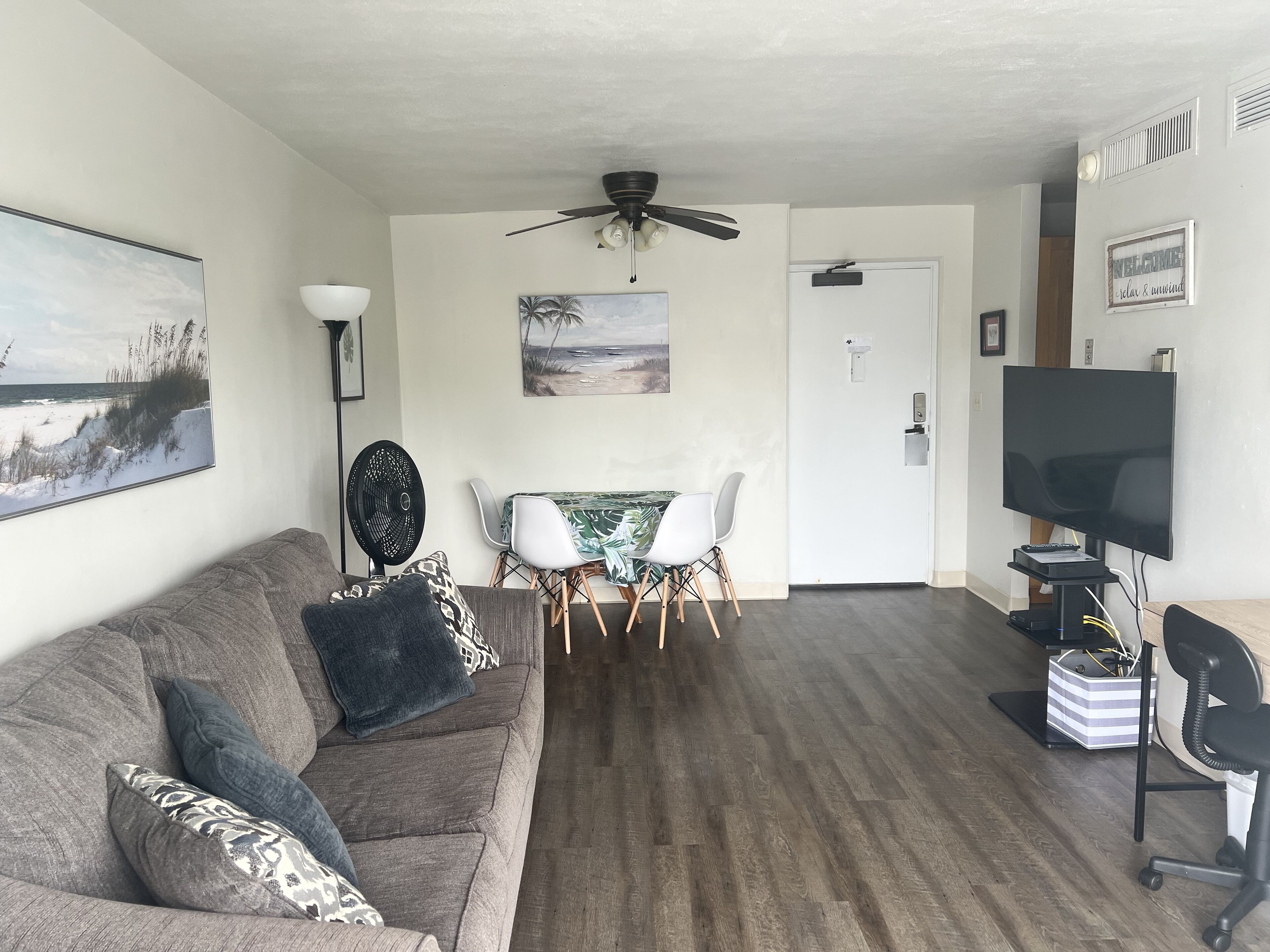 A living room with a gray couch, a dining table with chairs, a ceiling fan, a TV, and a painting on the wall.