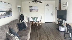 Uma sala de estar com sofá cinza, uma mesa de jantar com cadeiras, um ventilador de teto, uma televisão e um quadro na parede.