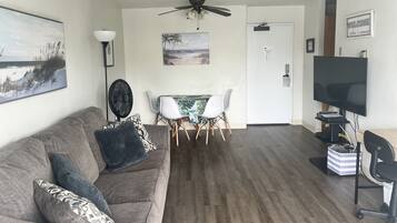 A living room with a gray couch, a dining table with chairs, a ceiling fan, a TV, and a painting on the wall.