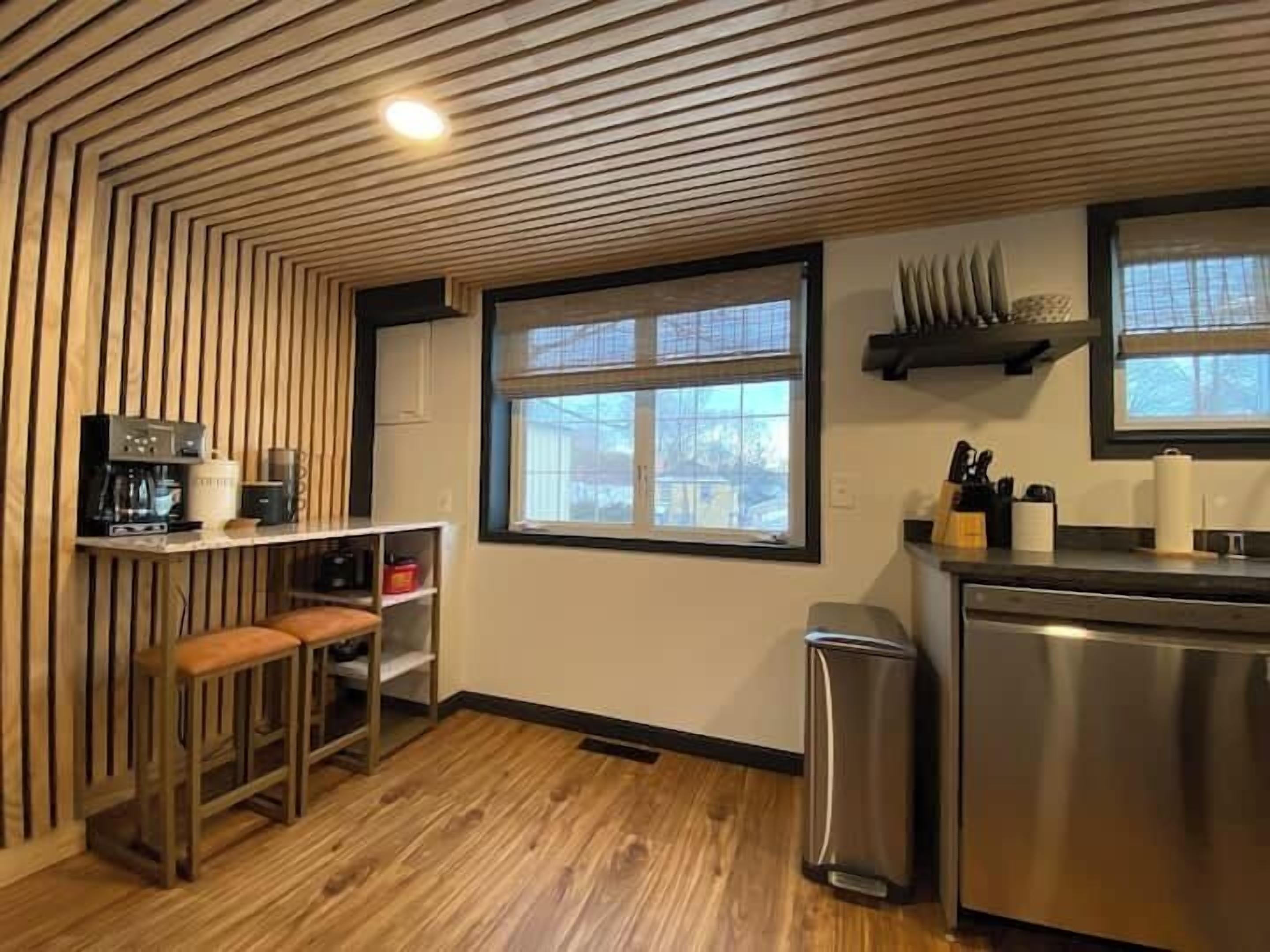 A modern kitchen with a wooden ceiling, a window with blinds, a stainless steel refrigerator, and a trash bin.