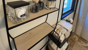 A bathroom with a mirror, shelves with toiletries, a window with blinds, and a towel rack. - Designer Home in Walkable Historic Greendale (Greendale)