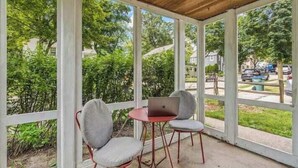 A covered outdoor seating area with two chairs and a table, overlooking a residential street and greenery. - Designer Home in Walkable Historic Greendale (Greendale)