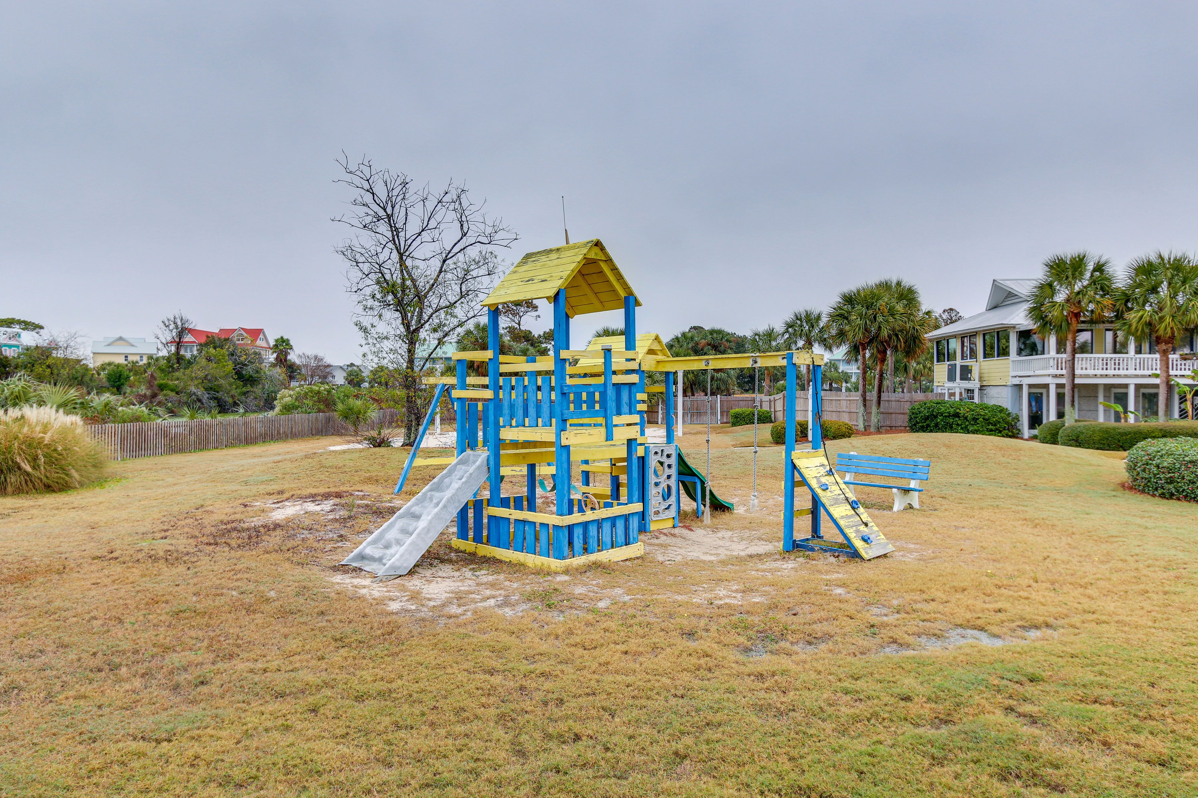 A playground with a blue and yellow play structure, a slide, and a swing set in a grassy area with houses in the background.