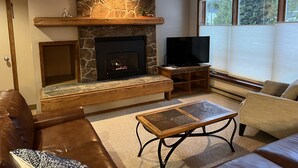 A cozy living room with a stone fireplace, a flat-screen TV, leather sofas, and a coffee table.