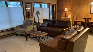 A living room with a brown leather sofa, a beige armchair, a wooden coffee table, and a dining area with a wooden table and chairs.