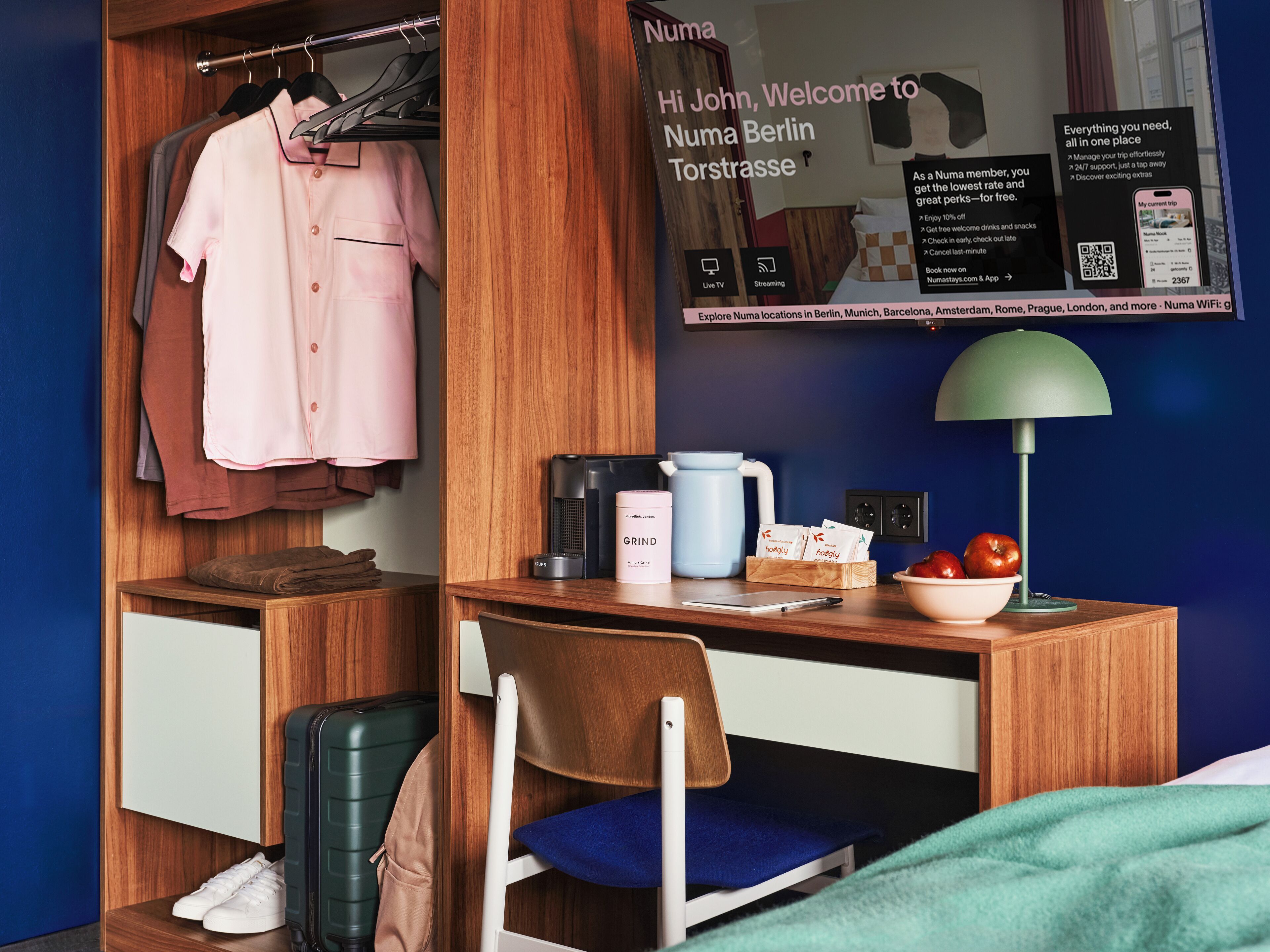 A compact hotel room with a wooden wardrobe, a desk with a laptop, a coffee maker, and a bowl of fruit.