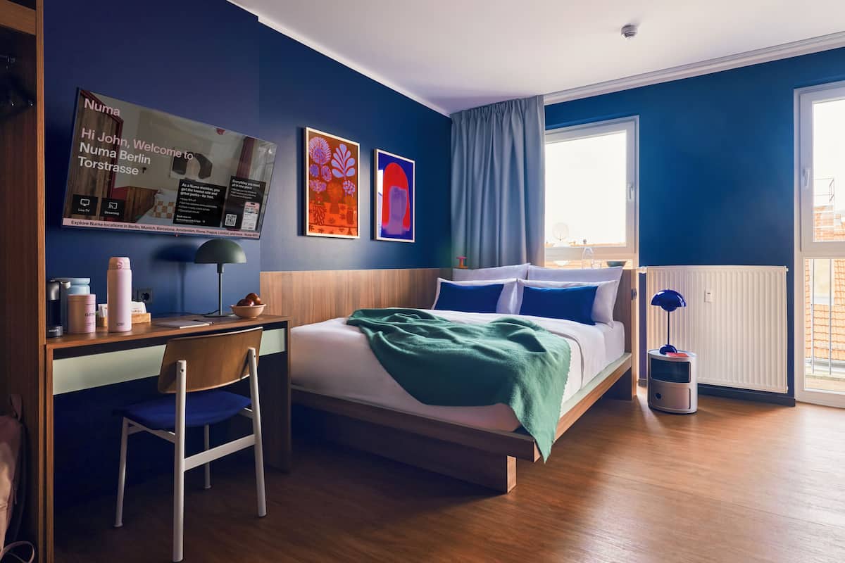 A modern hotel room with a bed, desk, and chair. There is a window with curtains, a radiator, and a wall-mounted TV.