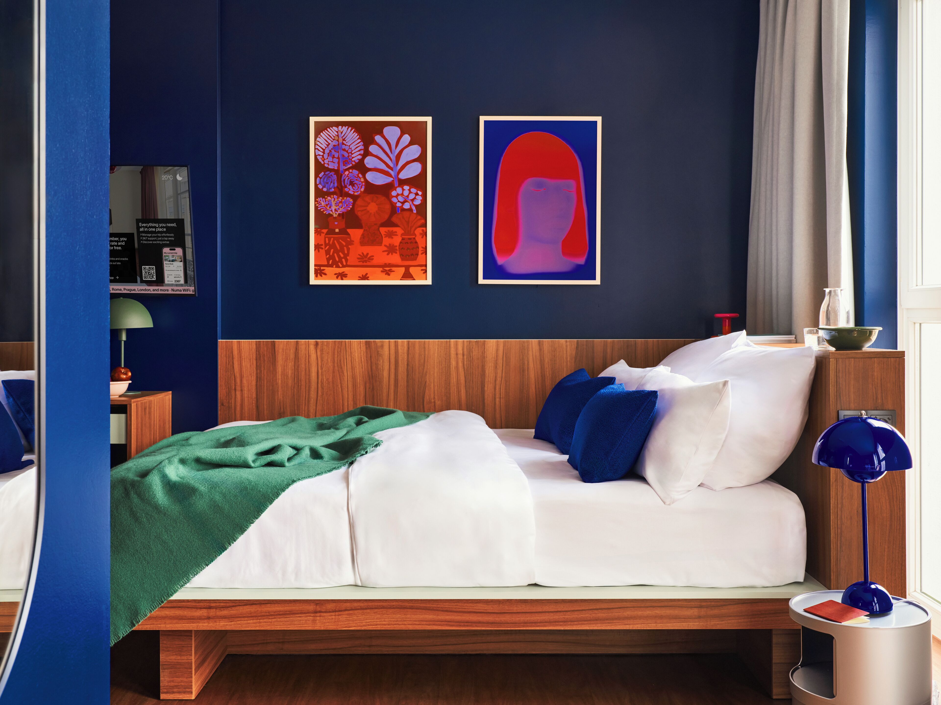 A hotel room with a bed, a nightstand, a lamp, and two framed artworks on the wall.