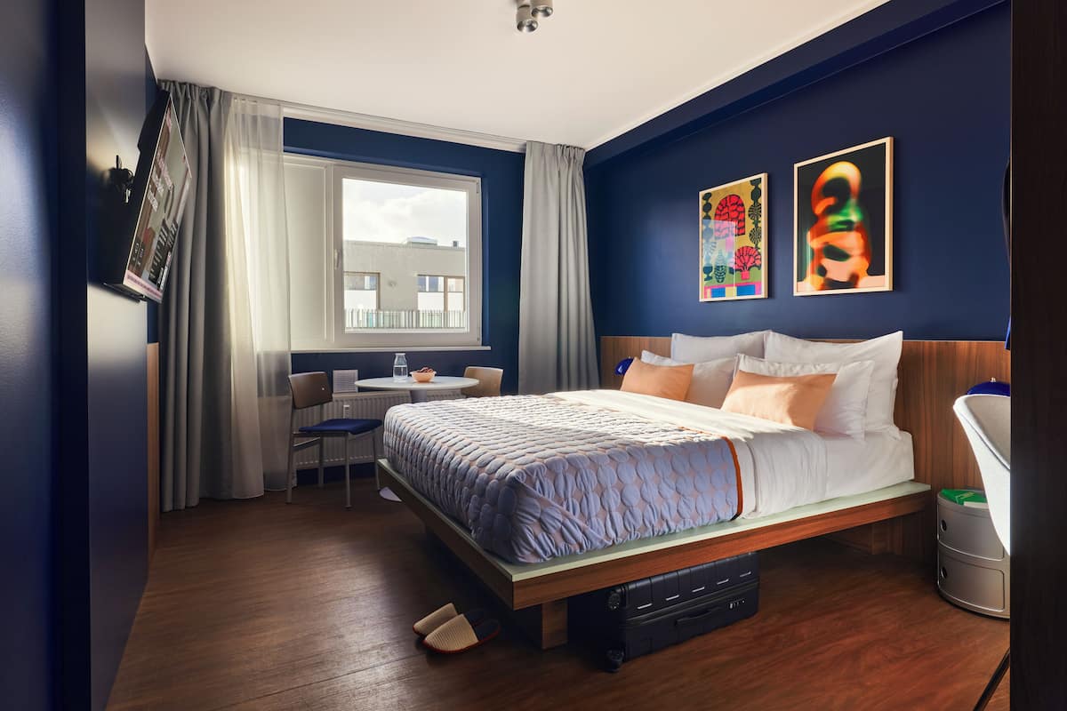 A modern hotel room with a large bed, a desk, and a window with a view of buildings.